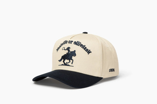 Front 3/4 view of a cream and dark blue baseball cap with "Nashville or Nashville" text arcing over an embroidered cowboy on a horse logo, plus "1806" on the side.