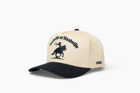 Front 3/4 view of a cream and dark blue baseball cap with "Nashville or Nashville" text arcing over an embroidered cowboy on a horse logo, plus "1806" on the side.